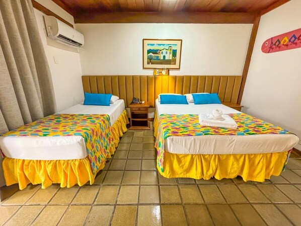 Habitación de hotel con dos camas, cada una con una colcha de colores y falda amarilla. Hay un aire acondicionado instalado en la pared y un cuadro enmarcado sobre las camas.
