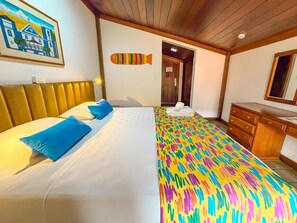 Une chambre d’hôtel avec un grand lit, une tête de lit en bois, une décoration murale colorée à rayures, une commode en bois et une porte donnant sur une autre pièce.