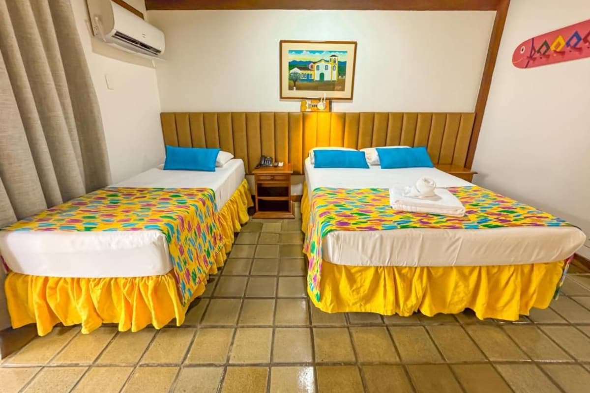 Habitación de hotel con dos camas, cada una con una colcha de colores y falda amarilla. Hay un aire acondicionado instalado en la pared y un cuadro enmarcado sobre las camas.