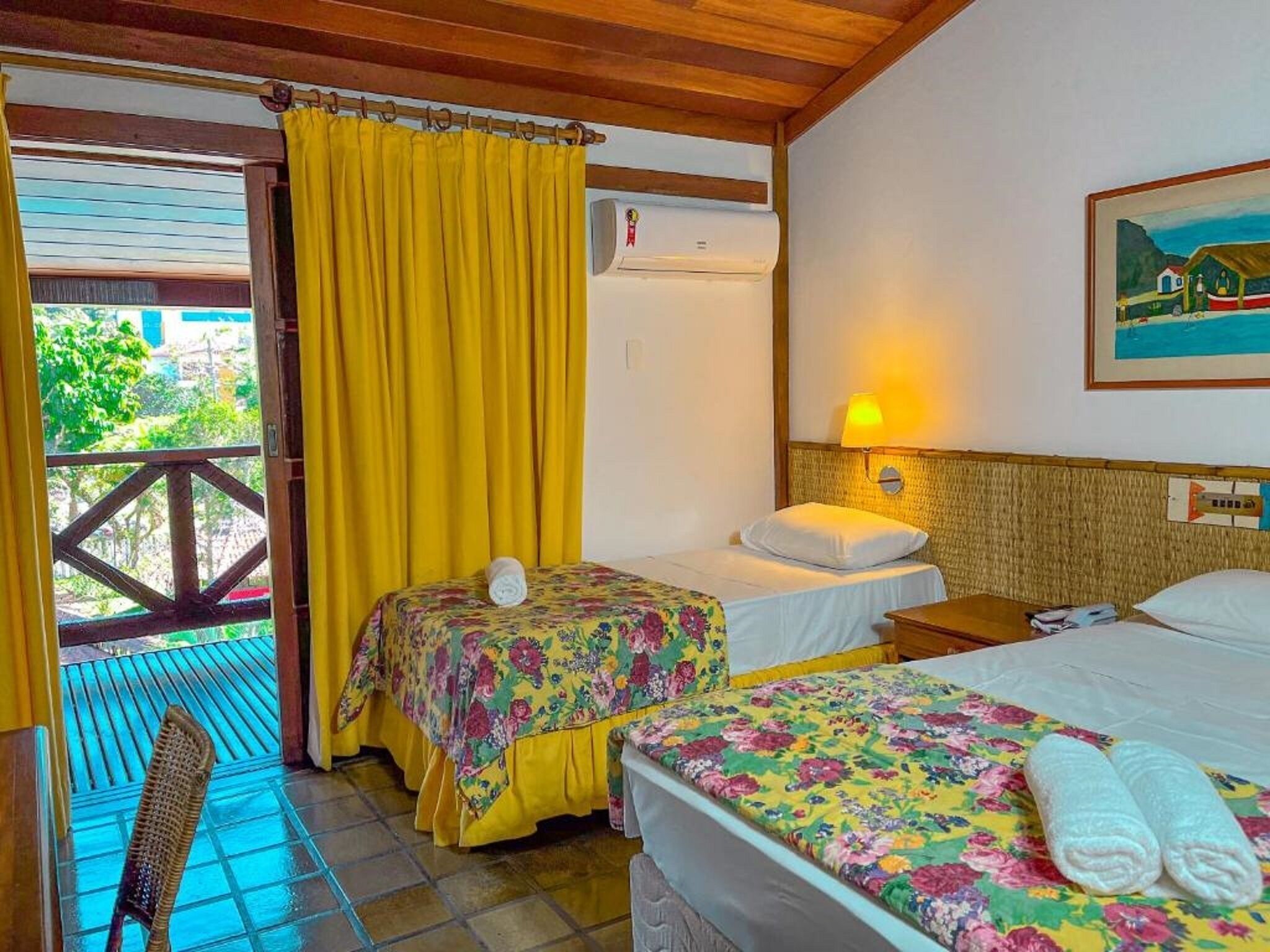 A room with two beds, a balcony with yellow curtains, a wicker chair, and a painting on the wall.