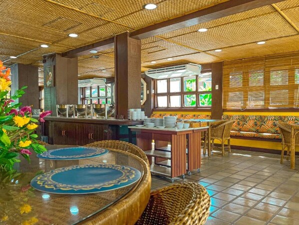 Ein Restaurant im tropischen Stil mit Bar, Sitzgelegenheiten mit gewobenen Stühlen und einer dekorativen Decke.