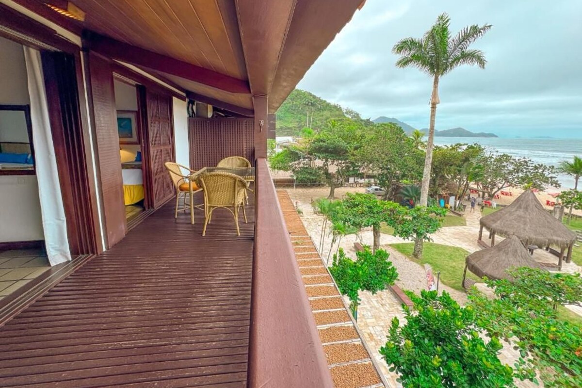Un balcón con piso de madera, sillas de mimbre y vistas a la playa y palmeras.