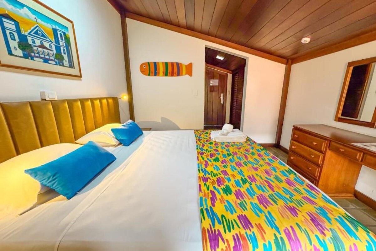 Habitación de hotel con una cama grande, un cabecero de madera, un adorno de pared con rayas de colores, un aparador de madera y una puerta que da a otra habitación.