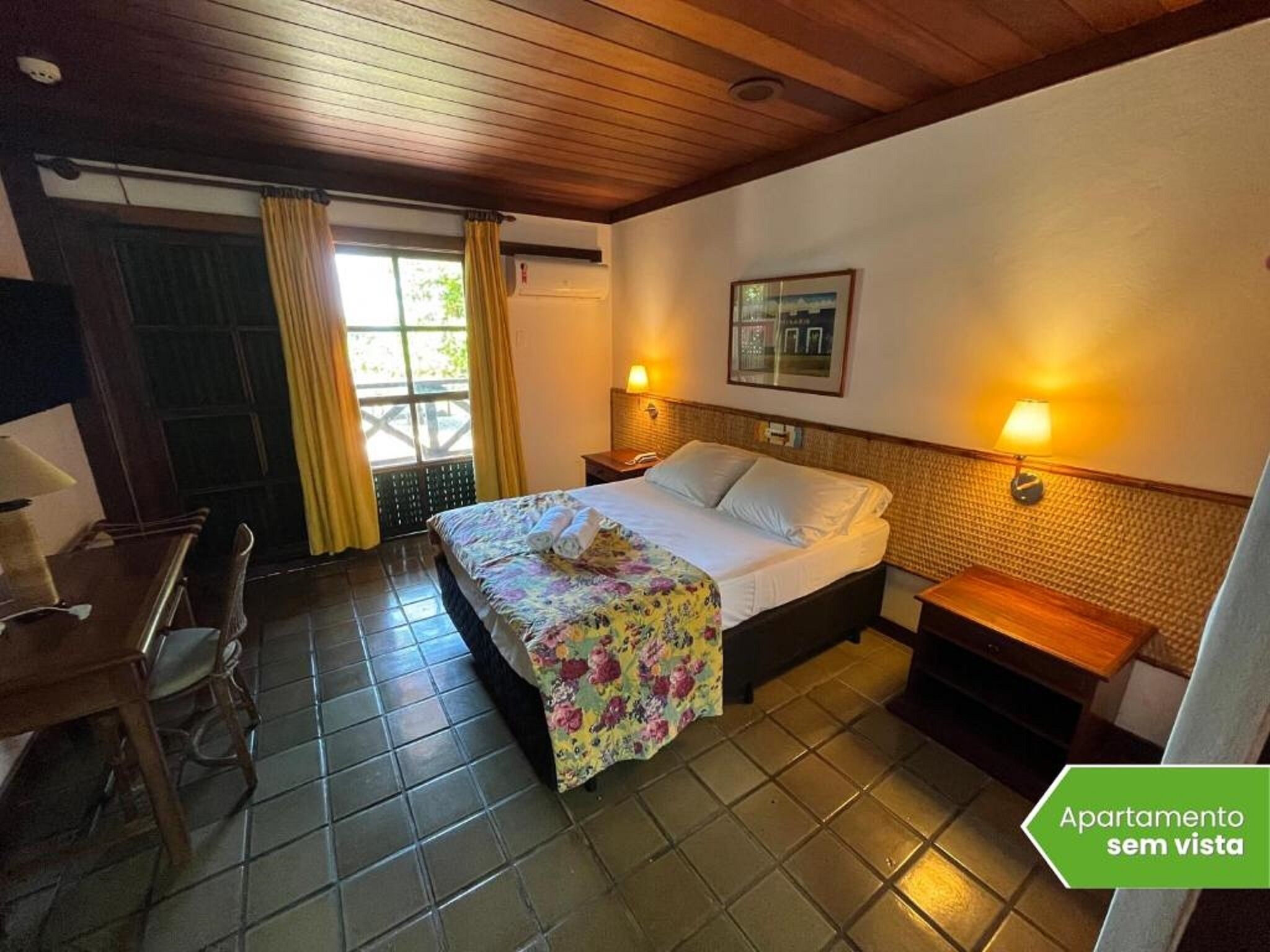 A hotel room with a bed, a desk, a chair, a window with curtains, a wall-mounted lamp, and a framed picture.