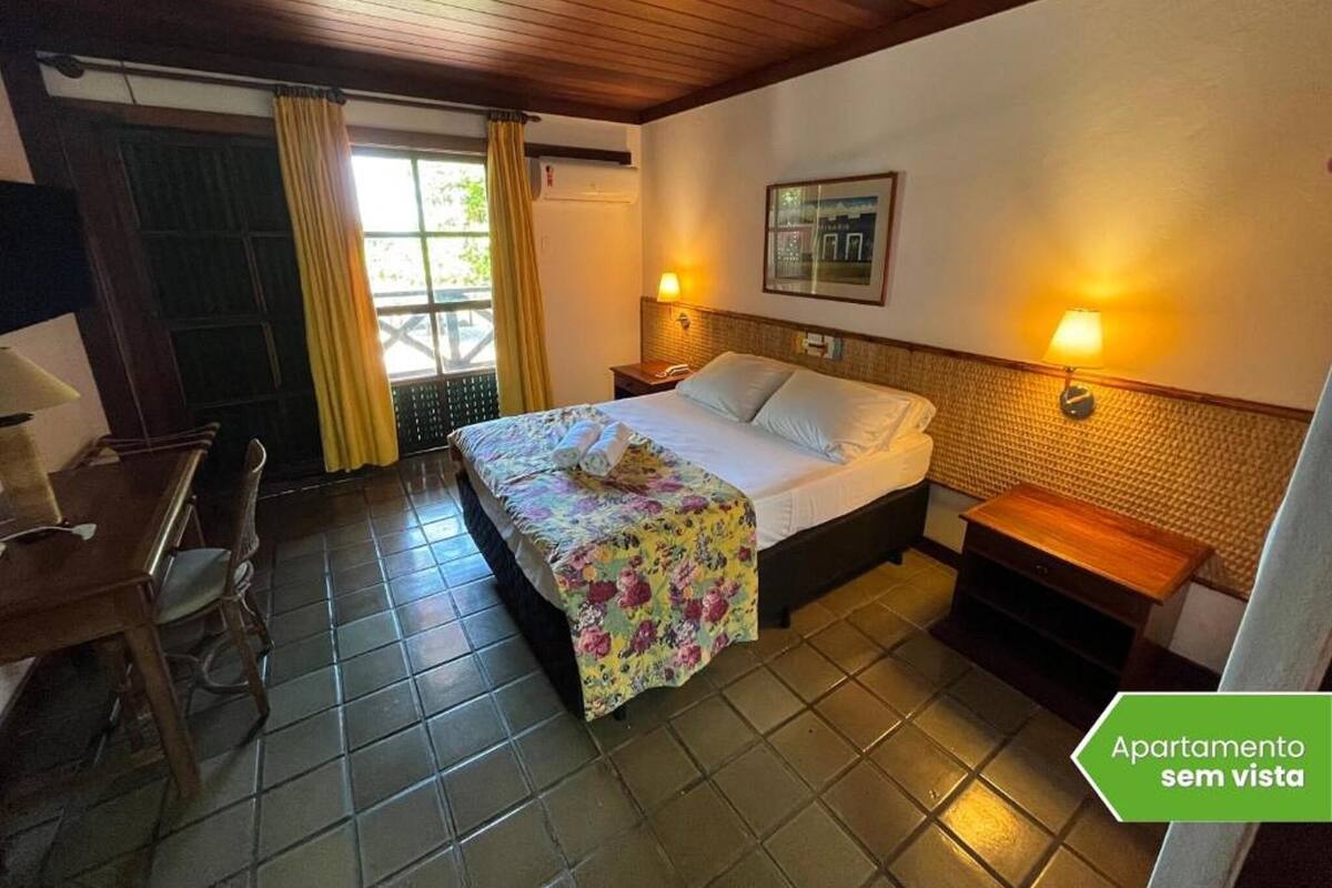 Une chambre d’hôtel comprenant un lit, un bureau, une chaise, une fenêtre avec des rideaux, une lampe fixée au mur et un tableau encadré.