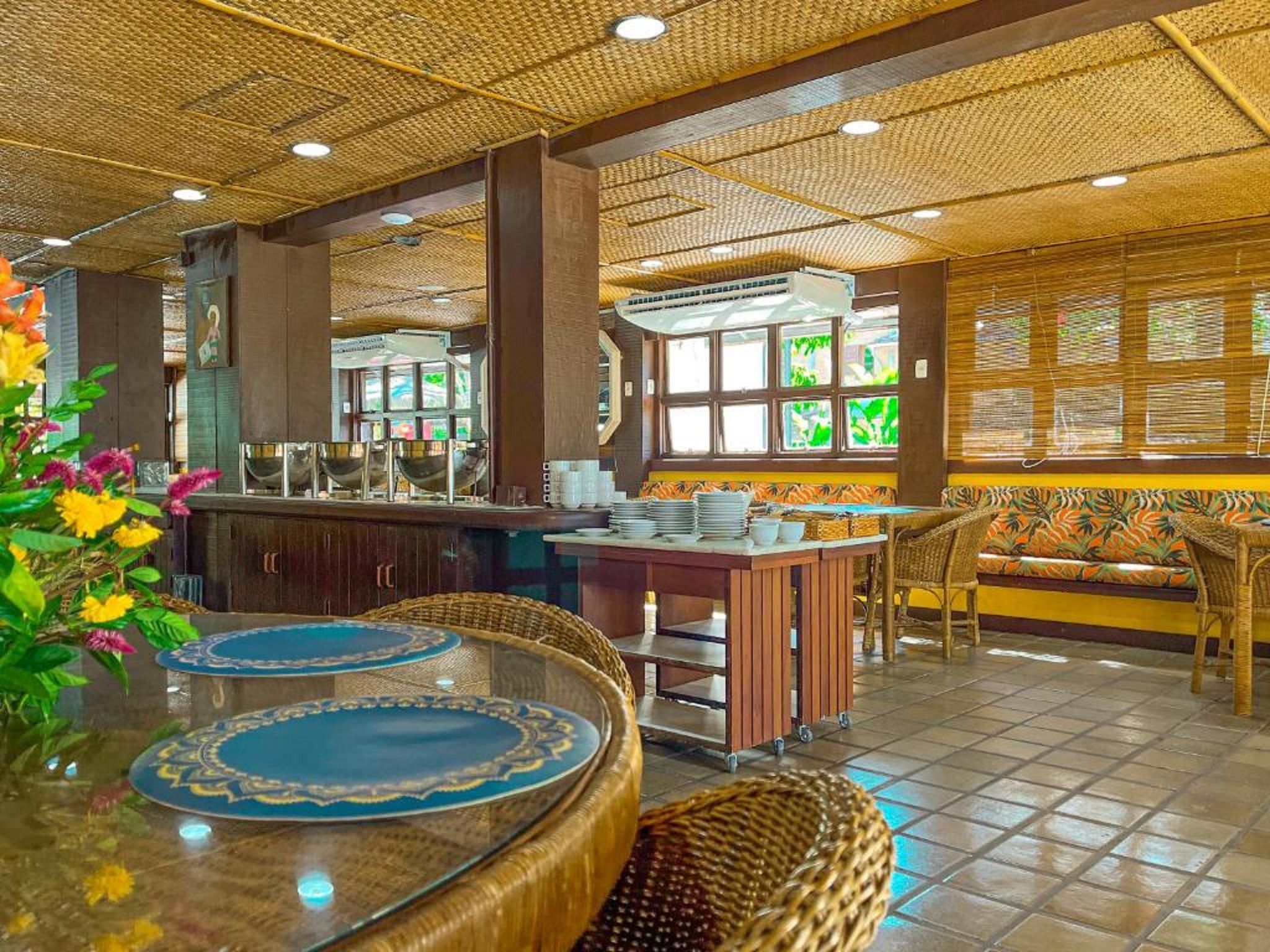 Een restaurant met een tropisch thema, met een bar, zitplaatsen met geweven stoelen en een decoratief plafond.