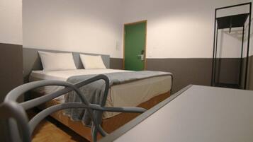 Uma cama de solteiro com cabeceira cinza, uma porta verde e uma divisória de vidro à direita.