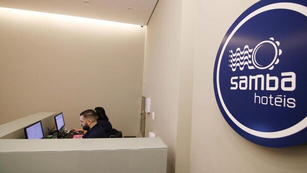 A reception desk with two monitors, a person seated behind it, and a logo on the wall reading "samba hotels". - Samba Belo Horizonte Vintage Apart Hotel (Belo Horizonte)