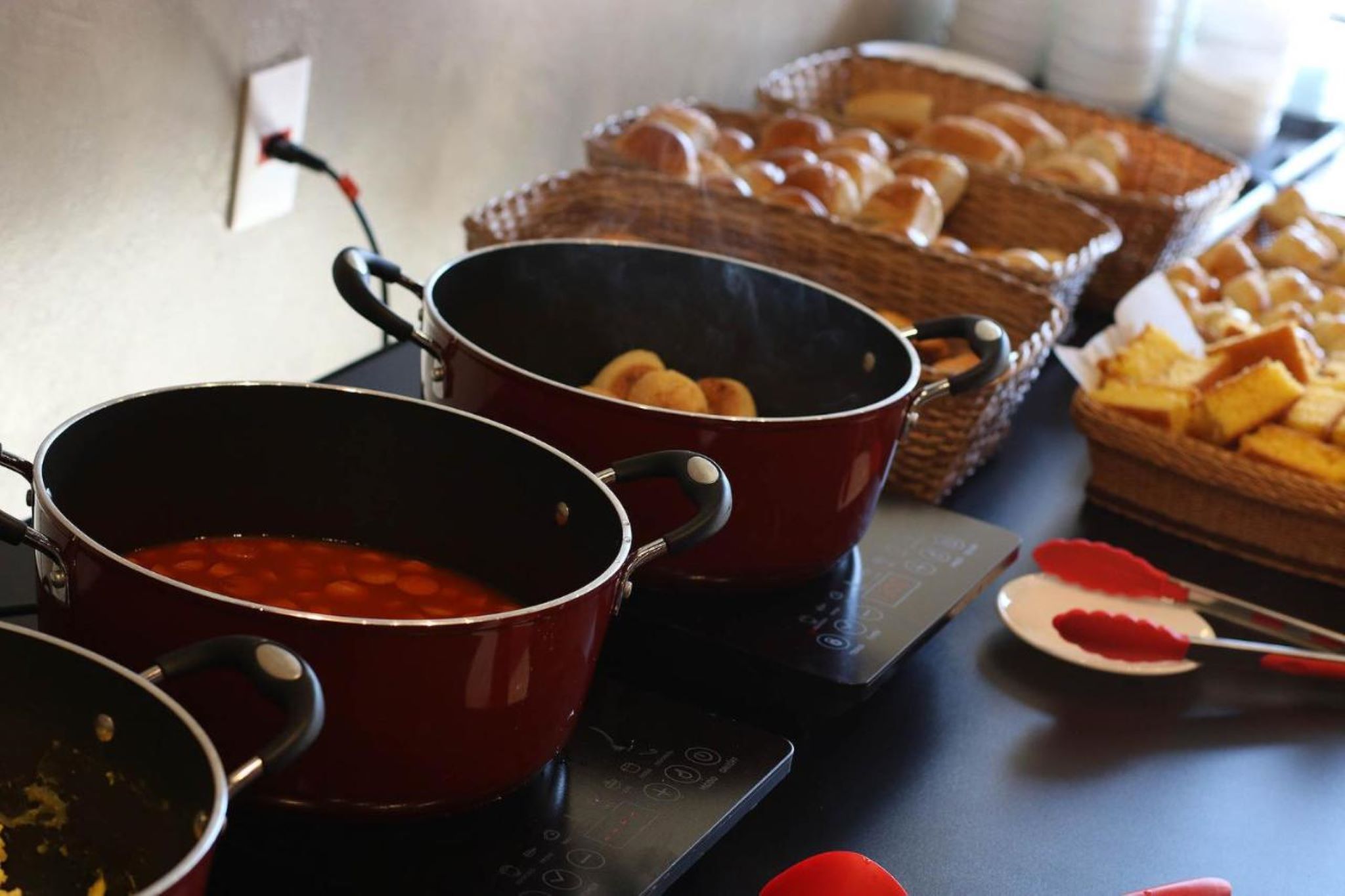 A kitchen counter with two red pots, a basket of bread rolls, and a plate with a red substance.