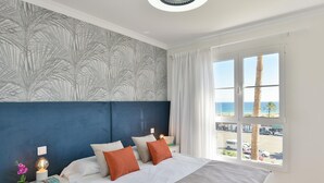 Ein Schlafzimmer mit Bett, Nachttischen, einer Lampe und Blick auf den Strand durch ein Fenster.
