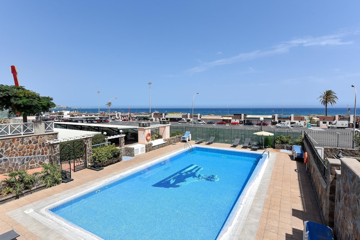 Une piscine avec vue sur la mer, entourée de chaises longues et d’un mur en pierre.
