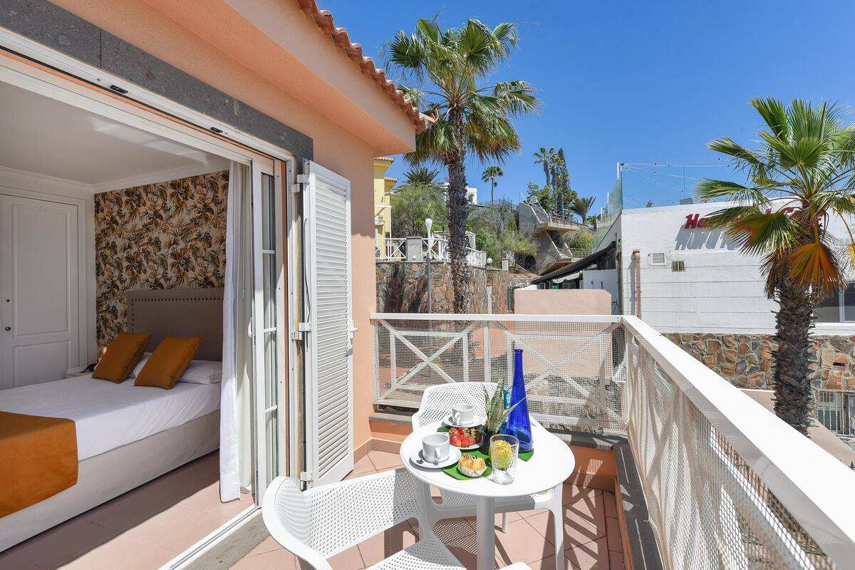 Un balcon avec un lit, une table avec une boisson, et une vue sur des palmiers et des immeubles.