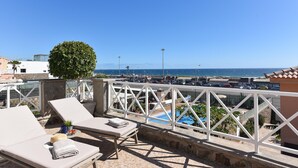 Eine Dachterrasse mit Liegestühlen, einem kleinen Baum und Blick auf das Meer sowie einen Parkplatz.