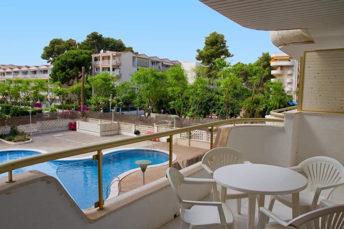 A balcony with a pool, white chairs, and a view of a residential area with trees and buildings.