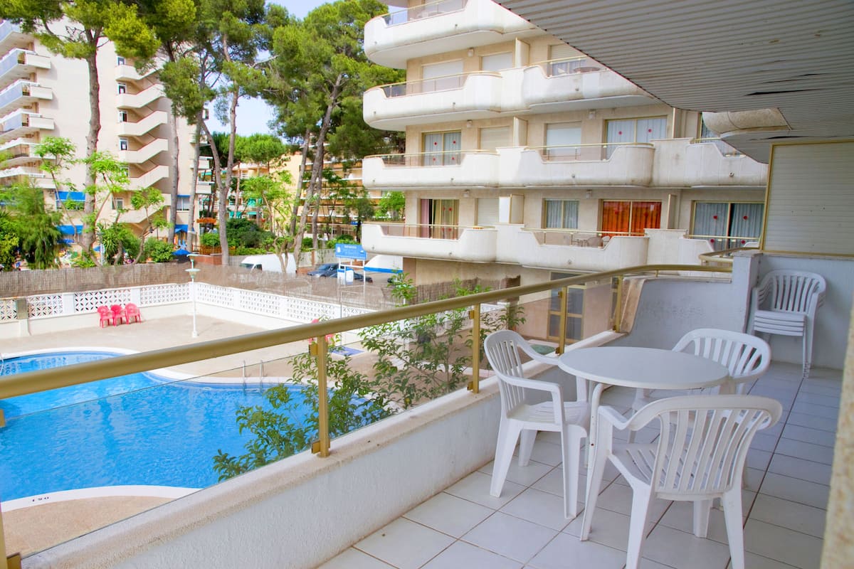 A balcony with a pool, white plastic chairs, and a view of a multi-story building with balconies.