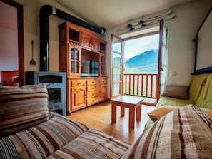 A cozy living room with a sofa, a TV, wooden cabinets, and a balcony with a mountain view. - Apartaments Pleta Bona (La Vall de Boi)