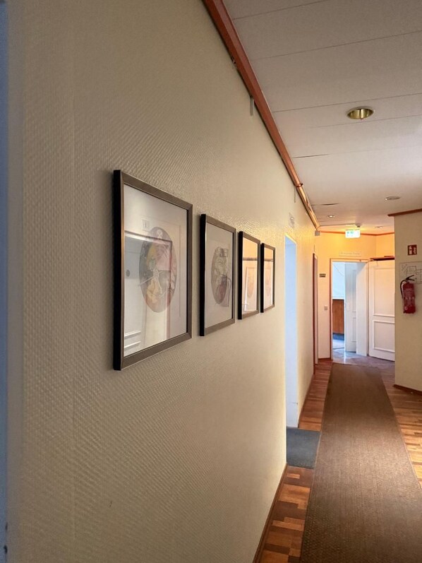 Ein Flur mit gerahmten Kunstwerken an der Wand, Holzdielen und einem Teppichbereich, der zu einer offenen Tür führt.