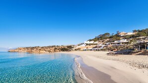 A beach with clear blue water, sandy shore, and buildings on a hillside. - Insotel Tarida Beach Resort & SPA (Sant Josep de sa Talaia)