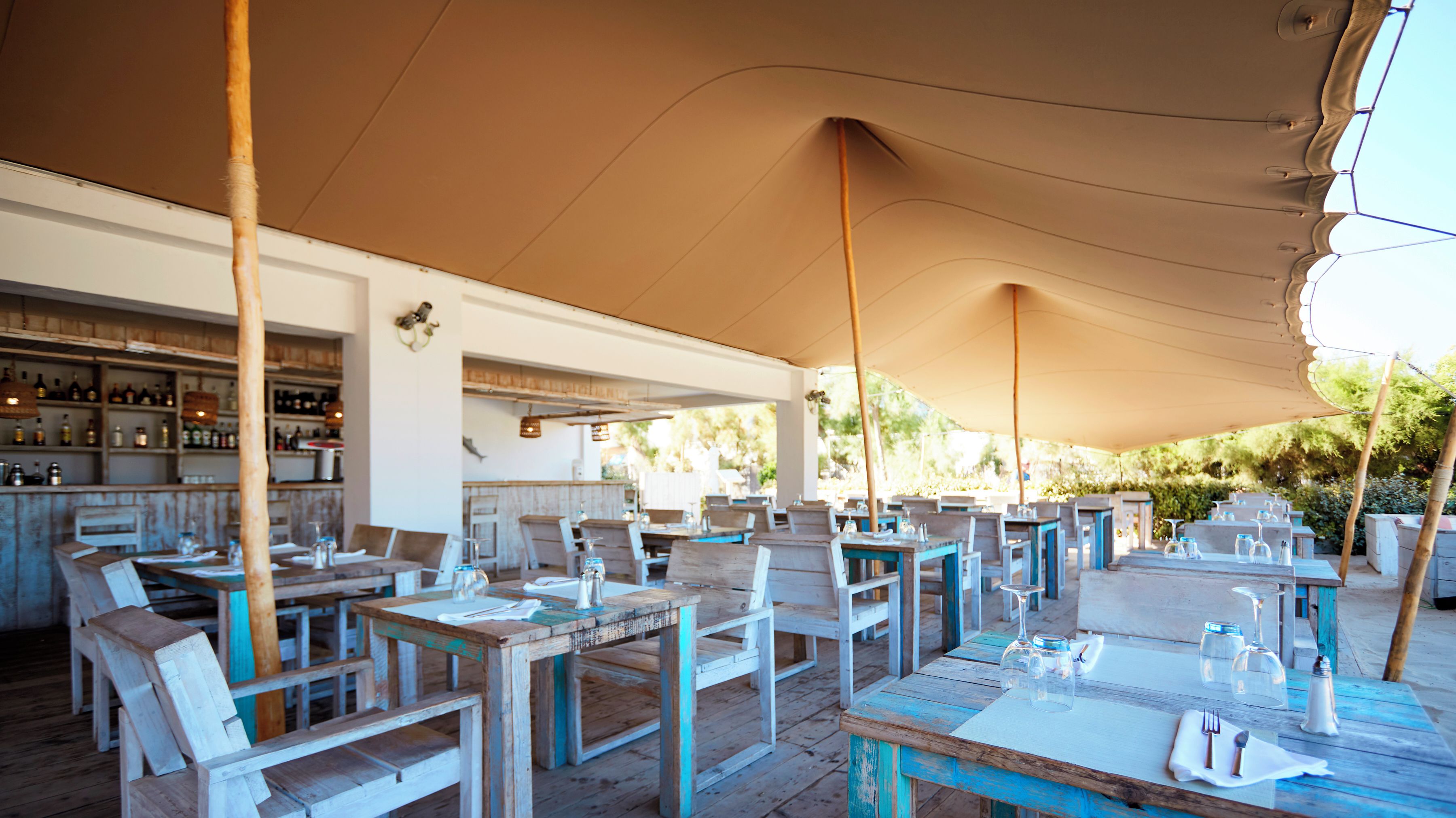 An outdoor dining area with wooden tables and chairs, a bar with shelves of bottles, and a covered section with a beige canopy.