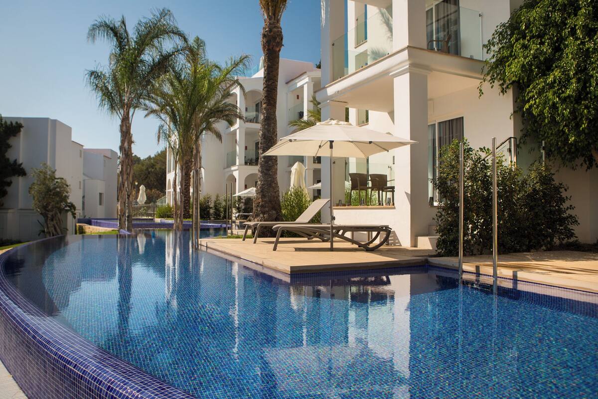 A swimming pool with lounge chairs and umbrellas, surrounded by palm trees and modern buildings.