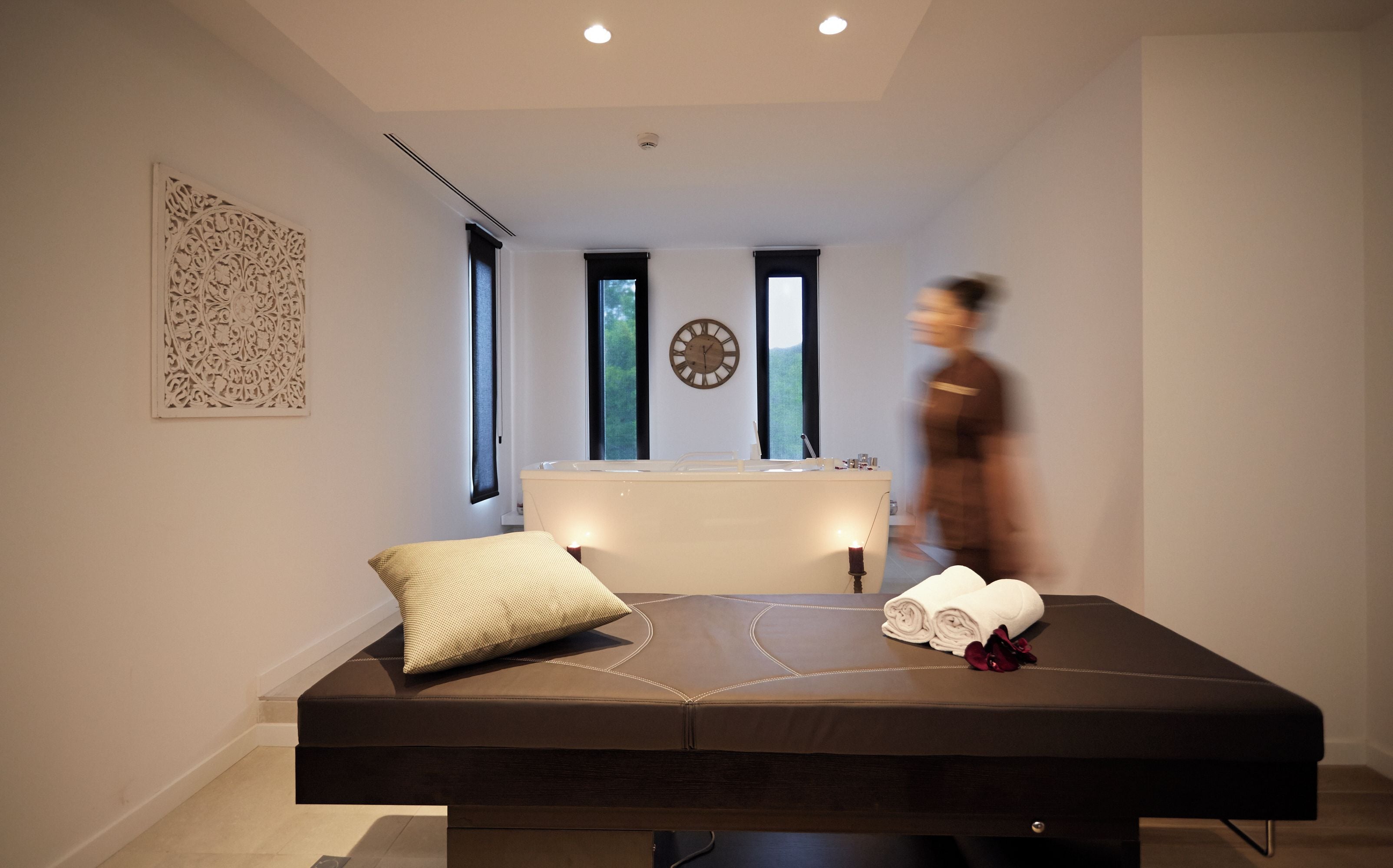 Una habitación moderna con una camilla de masaje, un reloj y una ventana que da a árboles.