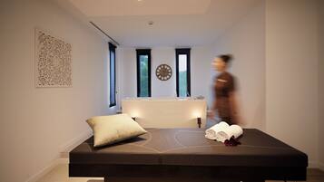 Una habitación moderna con una camilla de masaje, un reloj y una ventana que da a árboles.