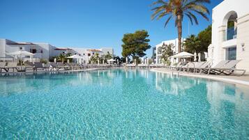 Una piscina cristalina con sillas de descanso y sombrillas, rodeada de edificios blancos y palmeras.