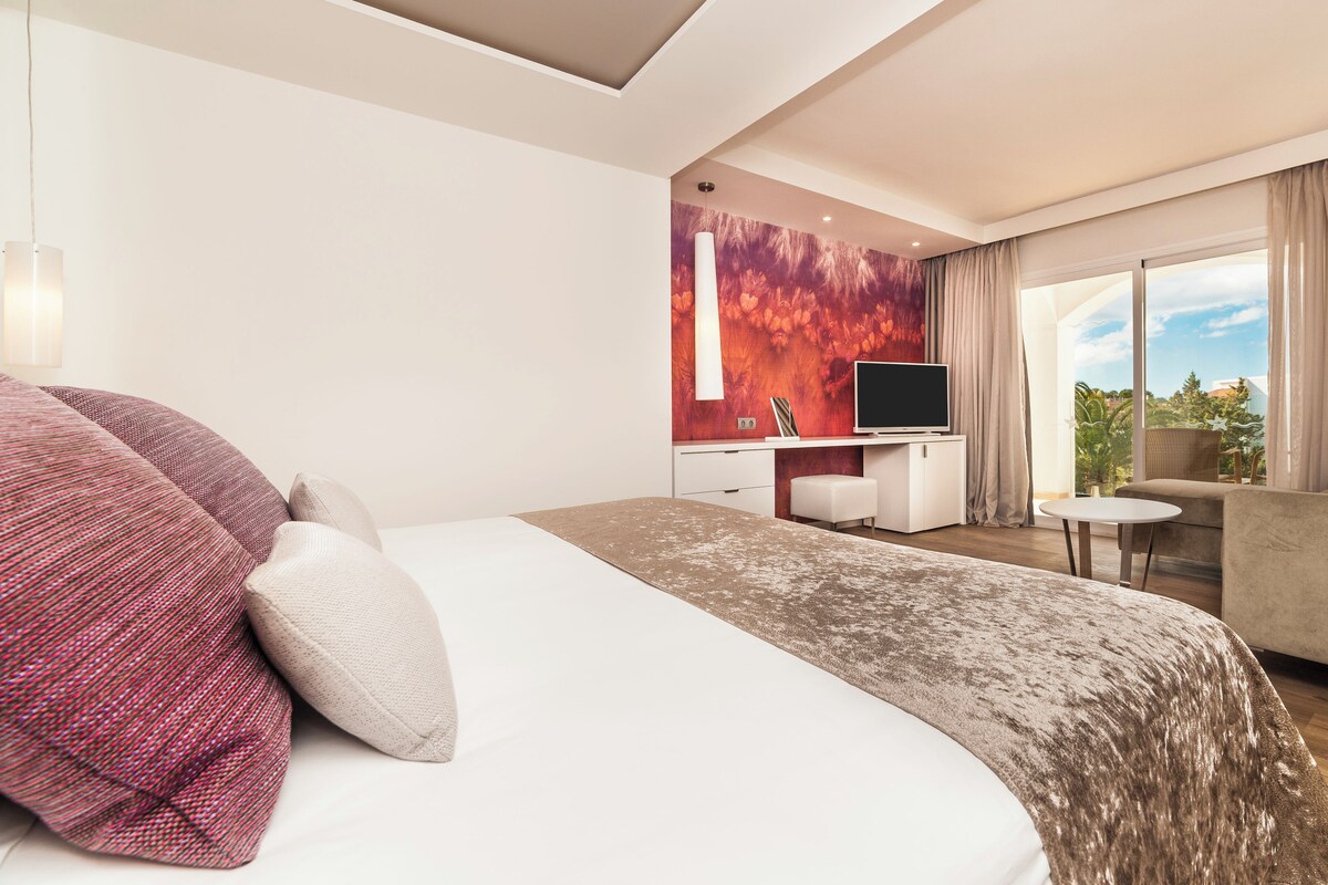 A modern hotel room with a large bed, a desk with a computer, a sofa, and a view of the outdoors.
