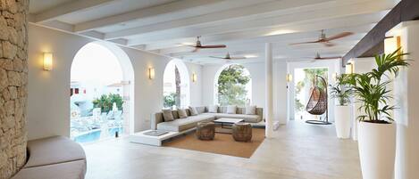 A spacious living area with a large sofa, a coffee table, and two side tables. There are ceiling fans, a stone wall, and potted plants.