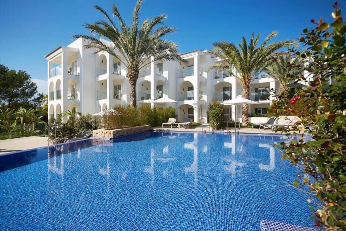 A modern hotel with a large swimming pool, palm trees, and a clear blue sky.