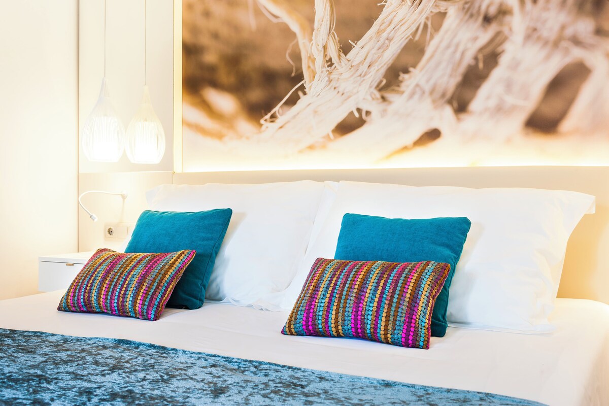 A bed with white linens, two teal pillows, and a colorful woven throw.