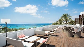Une terrasse sur le toit, avec des chaises et des tables en osier blanc, d’où l’on peut admirer une mer turquoise parsemée de bateaux.