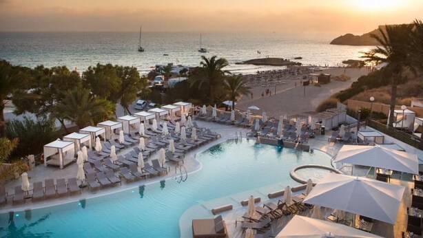A resort pool area with lounge chairs and umbrellas, a beach with people, and boats in the background. - Insotel Tarida Beach Resort & SPA (Sant Josep de sa Talaia)