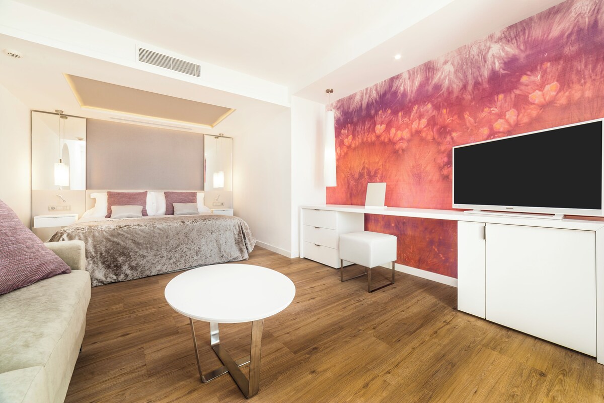A modern hotel room with a large bed, a white desk, a flat-screen TV, and a vibrant floral wall mural.