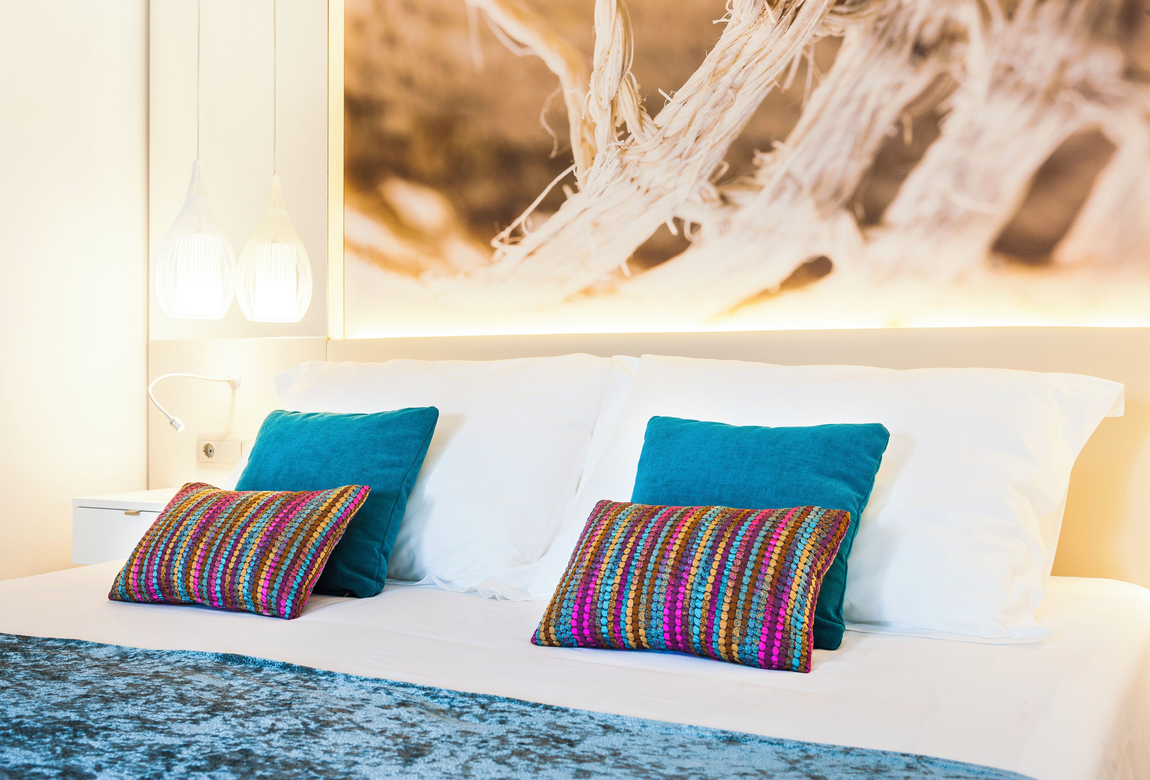 Una cama con sábanas blancas, dos almohadas turquesa y un colorido mantón tejido.