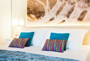 Un lit avec du linge de lit blanc, deux coussins turquoise et un plaid coloré en tissu tissé.