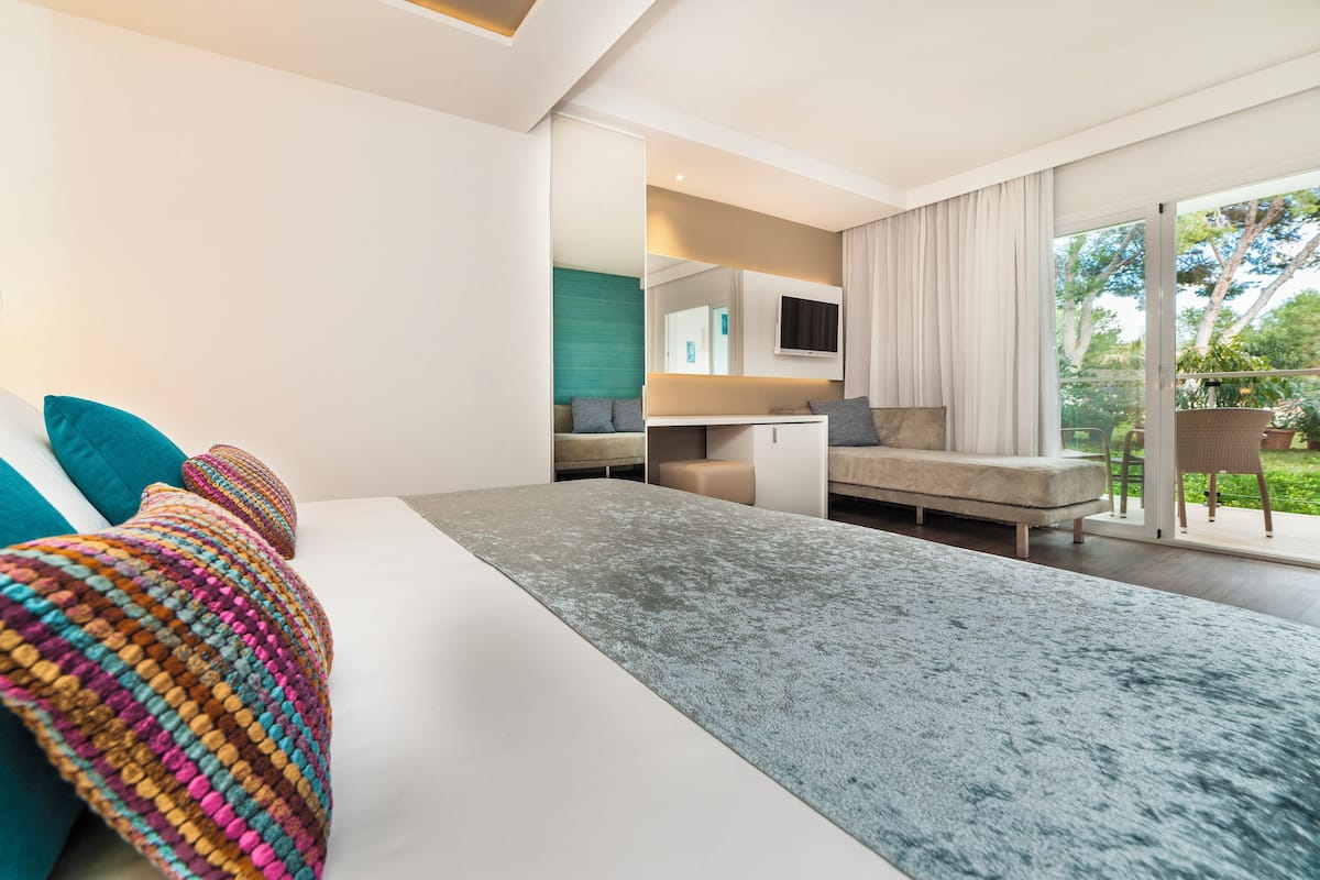 A modern bedroom with a large bed, a sofa, a TV, and a balcony with curtains.