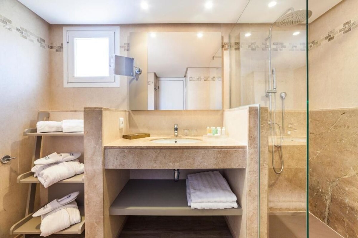 A modern bathroom with a glass-enclosed shower, a vanity with a sink, and a mirror. There are shelves with towels and toiletries.