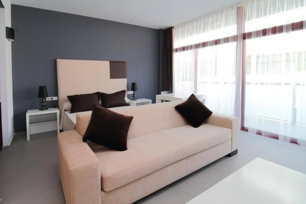 Une chambre d’hôtel moderne avec un grand lit, deux tables de chevet, un canapé et une grande fenêtre avec des rideaux.