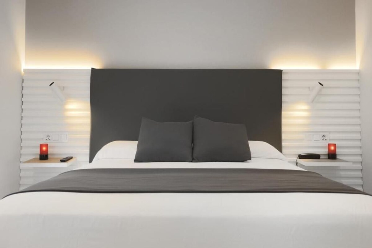 A modern bedroom with a large bed, dark headboard, bedside tables with red lamps, and a white wall with horizontal lines.