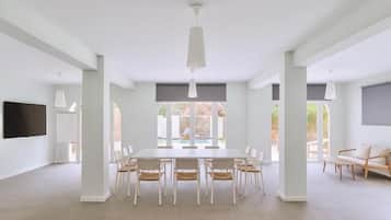 A spacious room with a large dining table surrounded by chairs, a flat-screen TV mounted on the wall, and large windows offering a view of the outdoors.