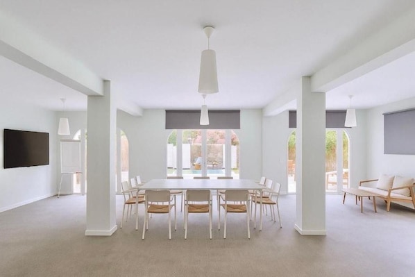 A spacious room with a large dining table surrounded by chairs, a flat-screen TV mounted on the wall, and large windows offering a view of the outdoors. - Sitges (Sitges)
