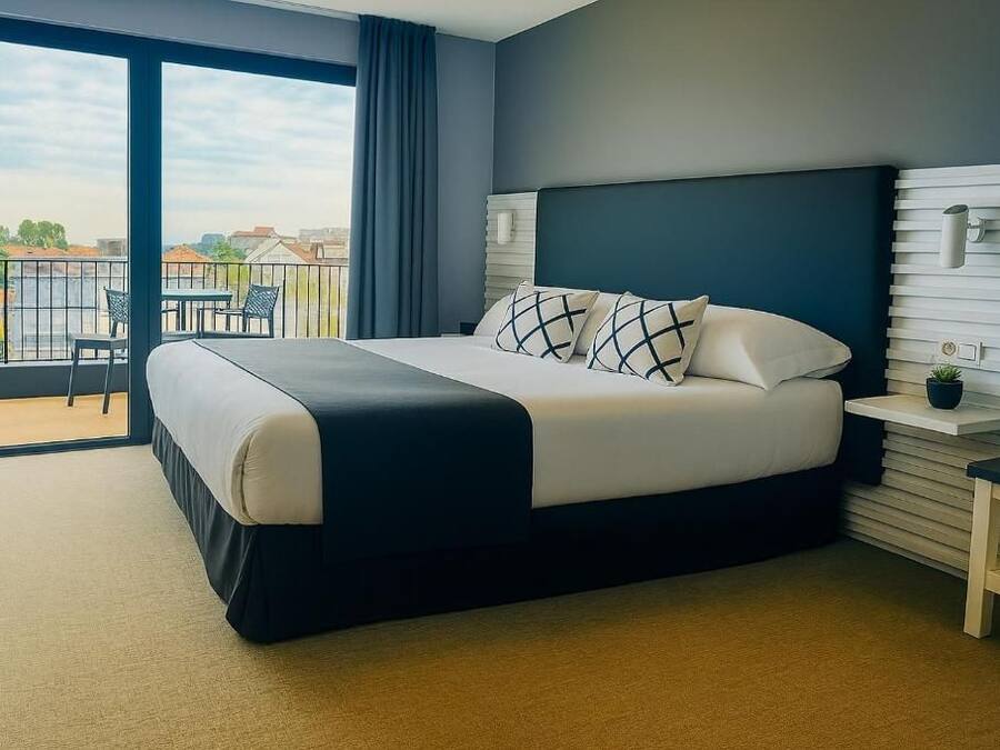 A modern hotel room with a large bed, a balcony with a view, and a small table with a plant.