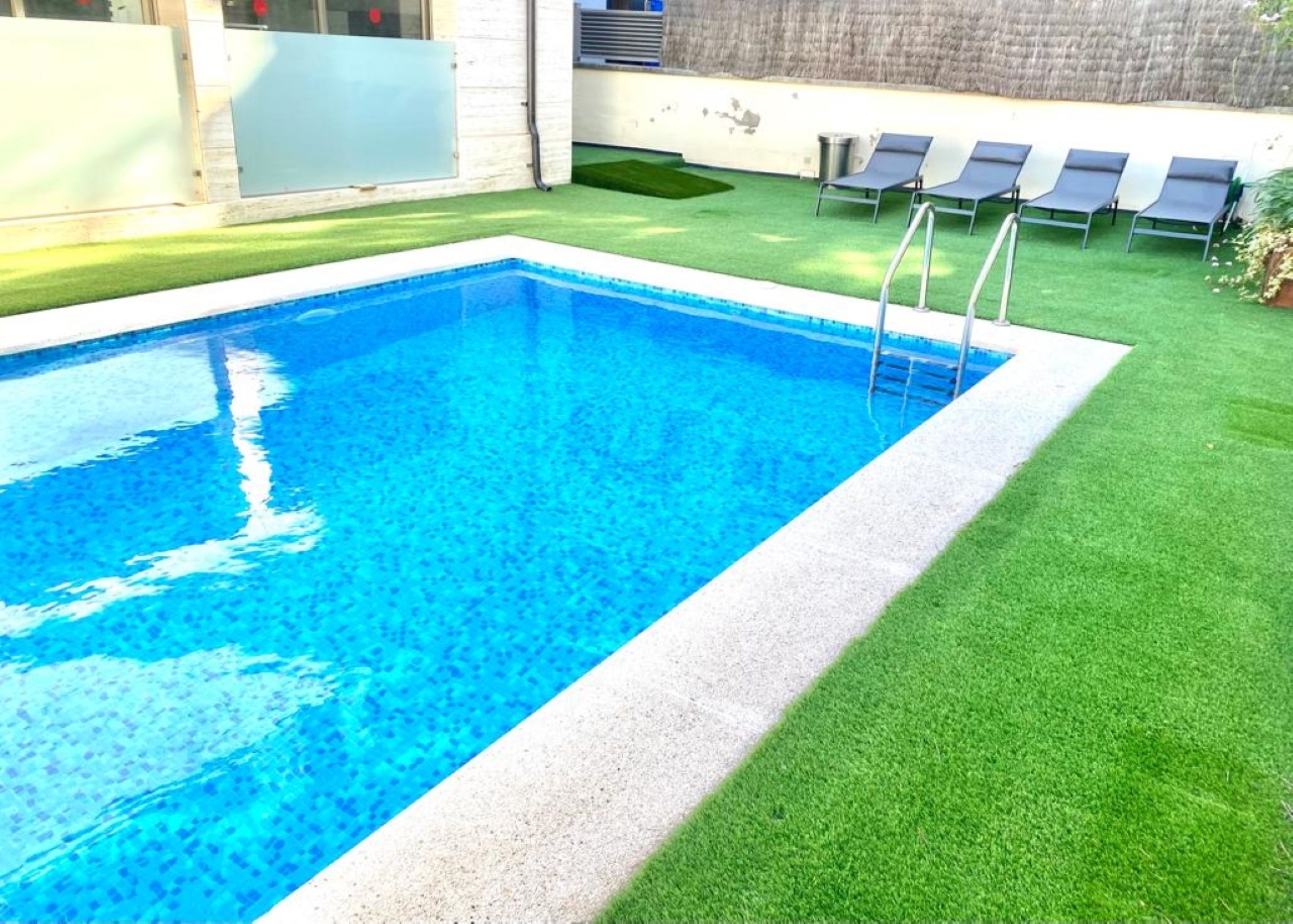 A rectangular swimming pool with a ladder, surrounded by artificial grass and a few lounge chairs.