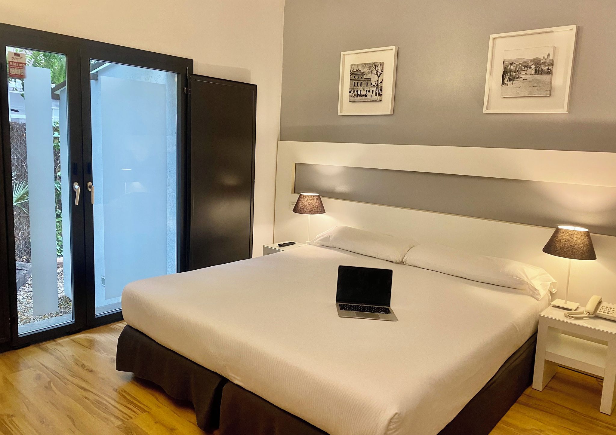 Ein Hotelzimmer mit einem großen Bett, einem Laptop auf dem Bett, zwei Nachttischlampen, einem Nachttisch mit einem Telefon und einer Tür zu einem Balkon.