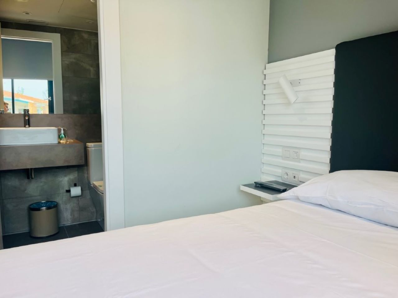A modern hotel room with a bed, a small bedside table, a nightstand, a mirror, and a bathroom with a sink and toilet.