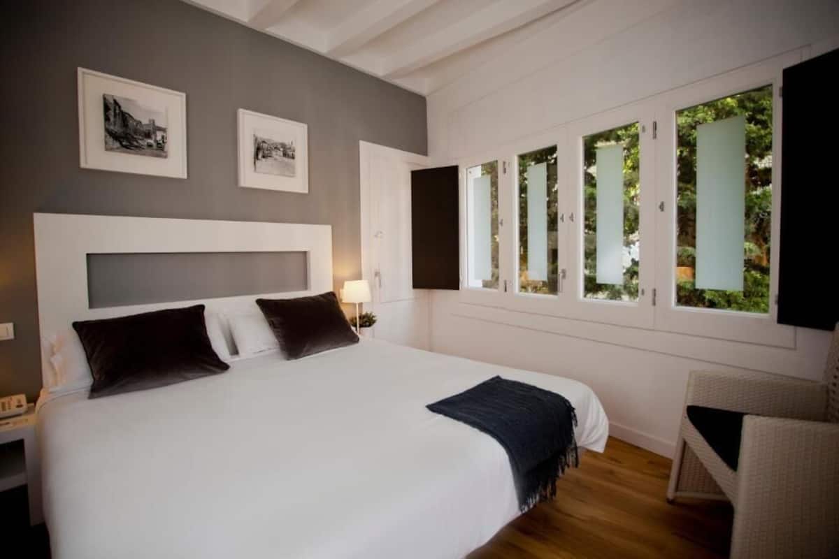 Un dormitorio con una cama grande, una mesita de noche, una lámpara y una silla. Hay cuadros enmarcados en la pared y una ventana con vistas a zonas verdes.