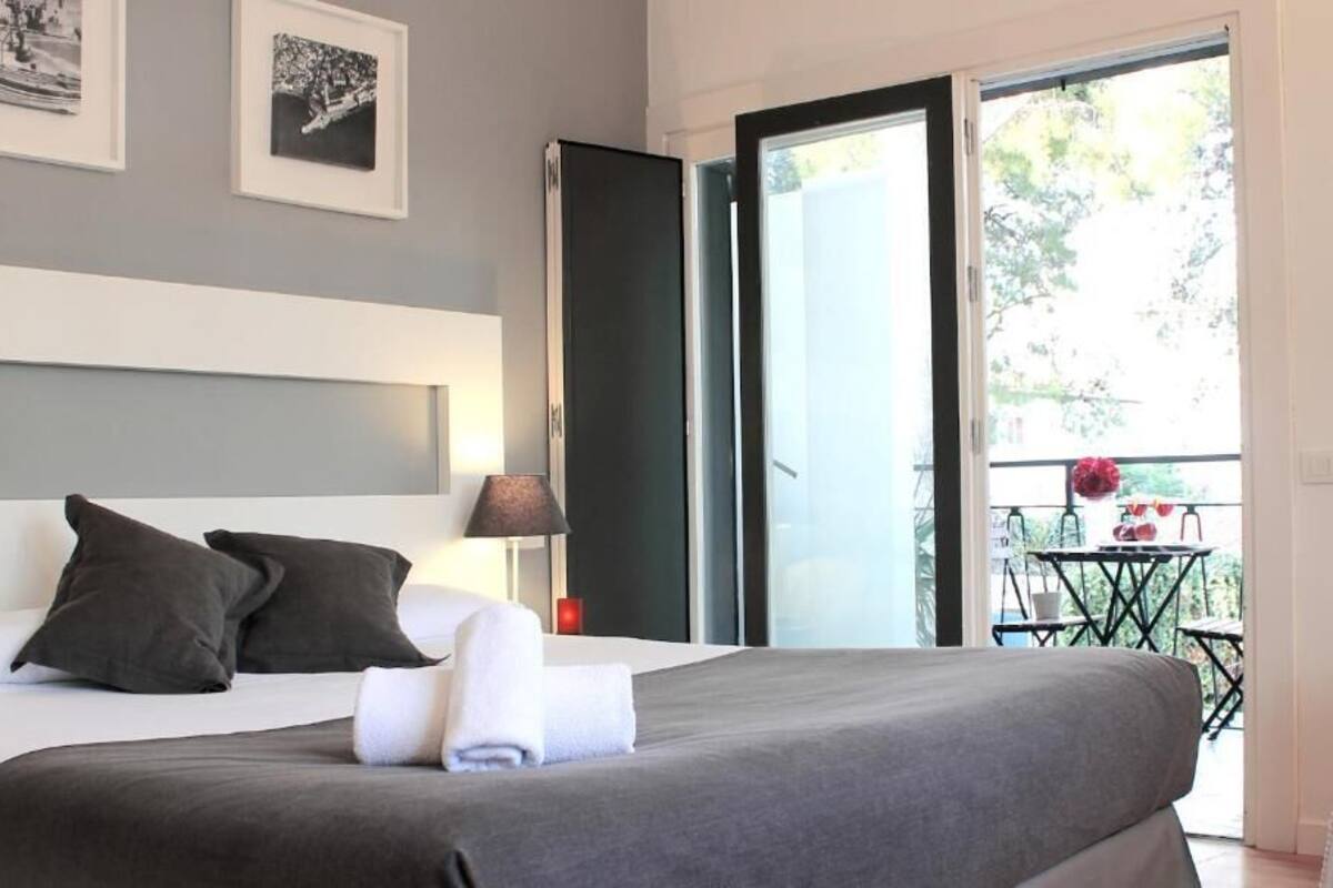 Una habitación de hotel moderna con una cama grande, una mesita de noche con lámpara y un balcón con mesa y sillas.