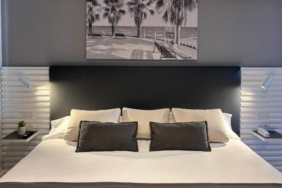 Un dormitorio moderno con una cama grande, un cabecero oscuro y una obra de arte con temática playera sobre ella.