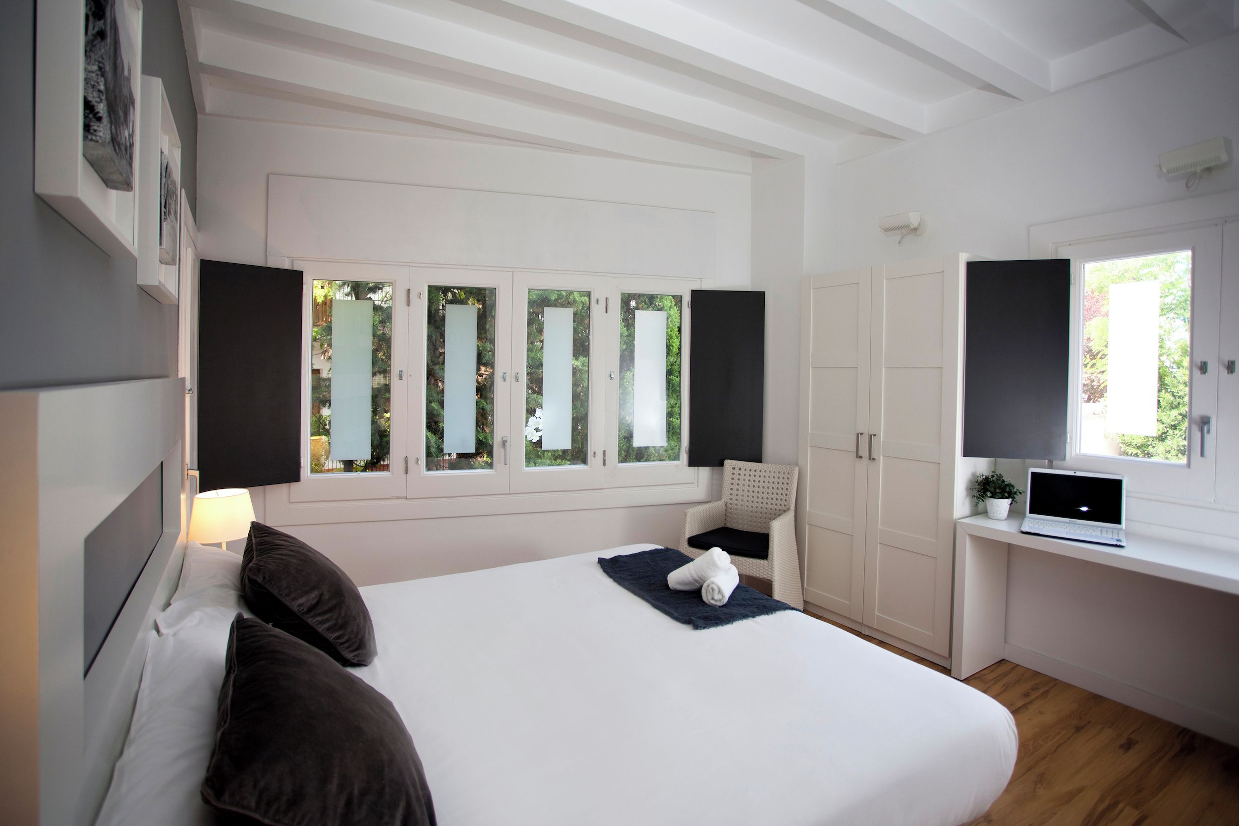 Habitación de hotel moderna con una cama grande, un escritorio con una computadora portátil, una ventana con vistas a zonas verdes y un armario blanco.